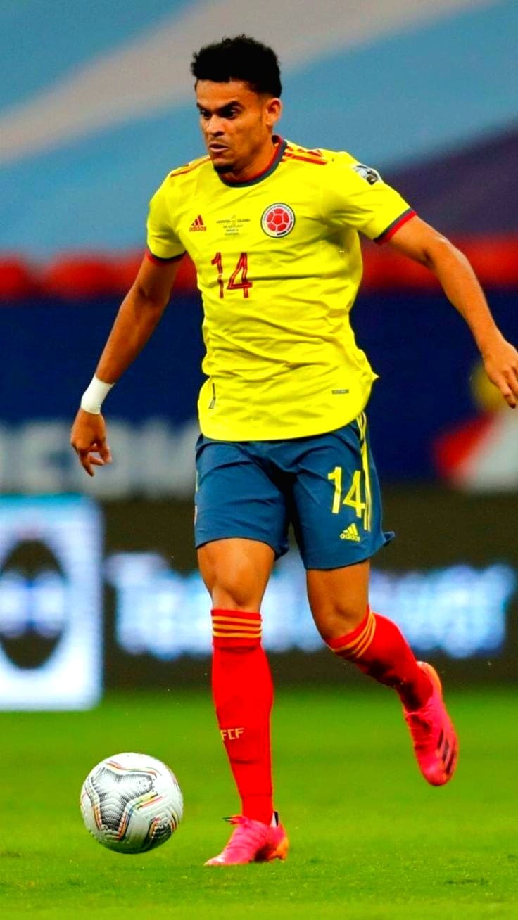 A soccer player in a yellow Colombia national team jersey with the number 14 dribbles a ball on the field during a match, as seen on Bulgaria IPTV. He sports blue shorts, red socks, and pink cleats.
